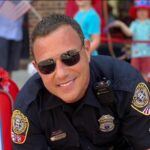 Police officer smiling in uniform, showcasing professionalism and approachability, with children and festive decorations in the background, relevant to community engagement and service.