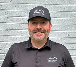 Justin Morris wearing a BisonWorks cap and shirt, smiling in front of a light gray brick wall, representing BisonWorks Home Improvement expertise and commitment to home projects.