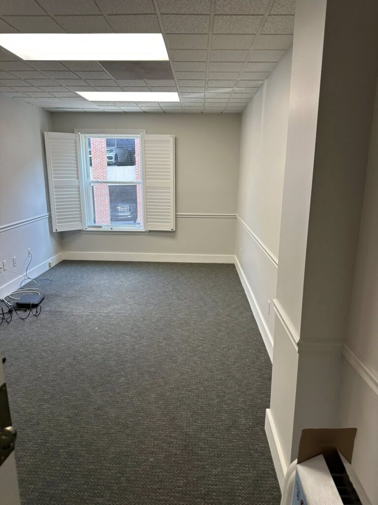 Empty office space with gray carpet, white walls, and a large window with shutters, showcasing potential for a home improvement project.
