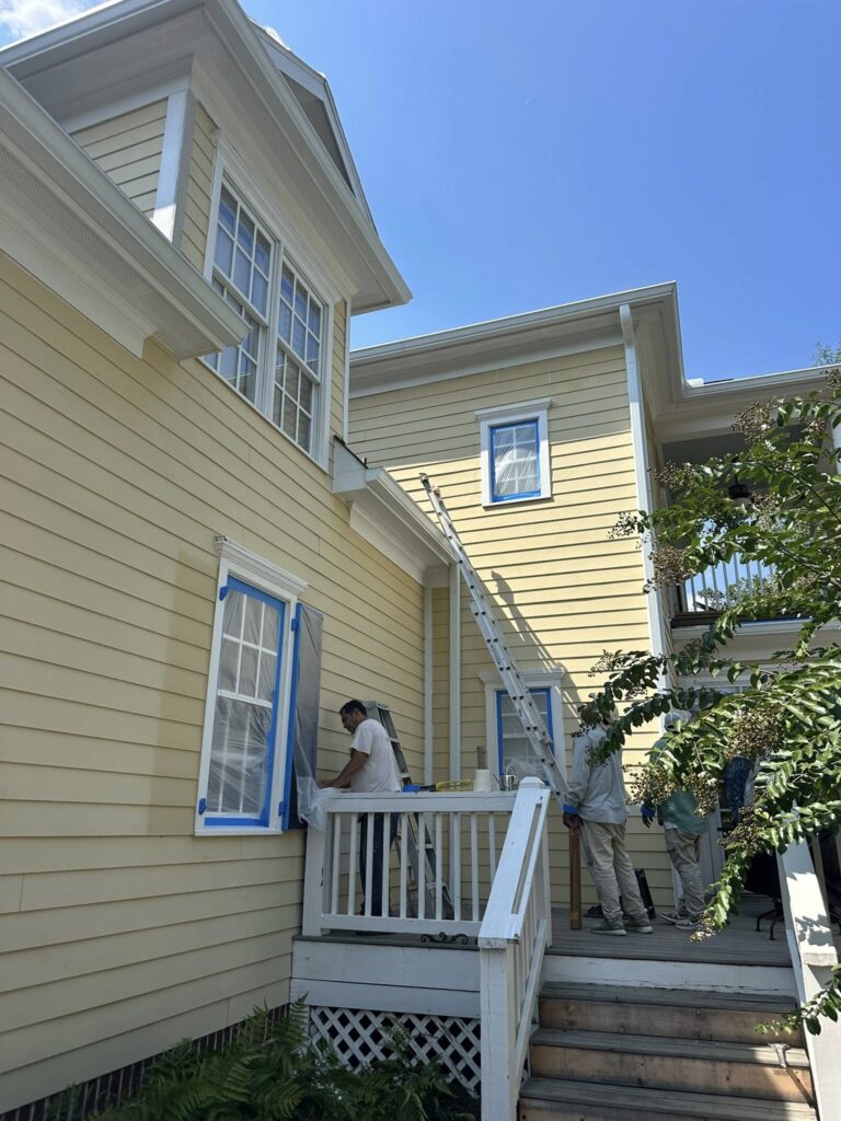Workers preparing a home for roofing installation, featuring a ladder, blue-taped windows, and a yellow exterior, emphasizing BisonWorks' commitment to quality home improvement services.