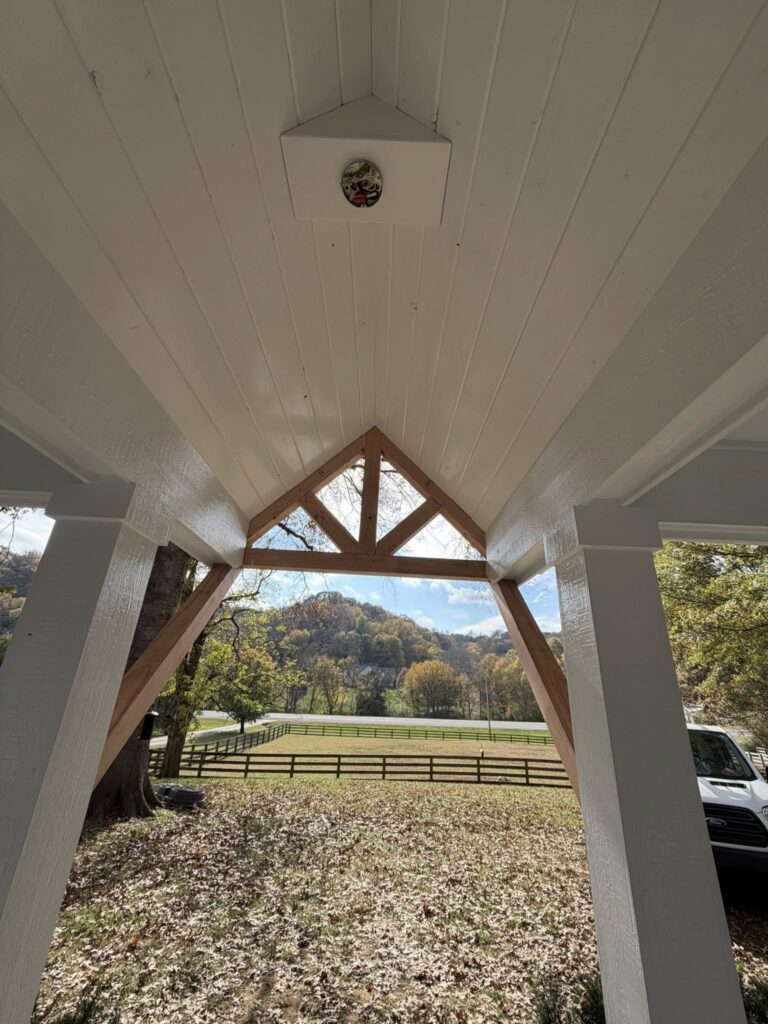 View from beneath a wooden roof structure, showcasing a white ceiling and triangular wooden beams, with a scenic landscape featuring trees and a fence in the background, emphasizing quality craftsmanship in home improvement.