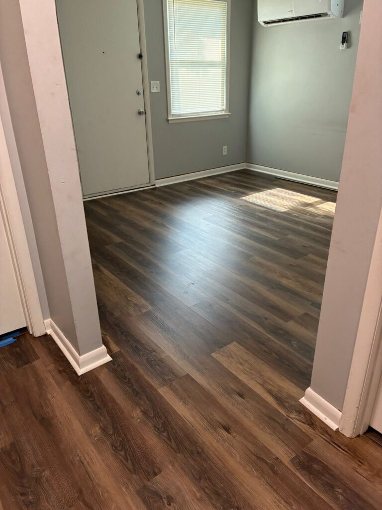 Newly installed wood laminate flooring in a modern, empty room with a front door, window, and air conditioning unit, showcasing a clean and inviting space.