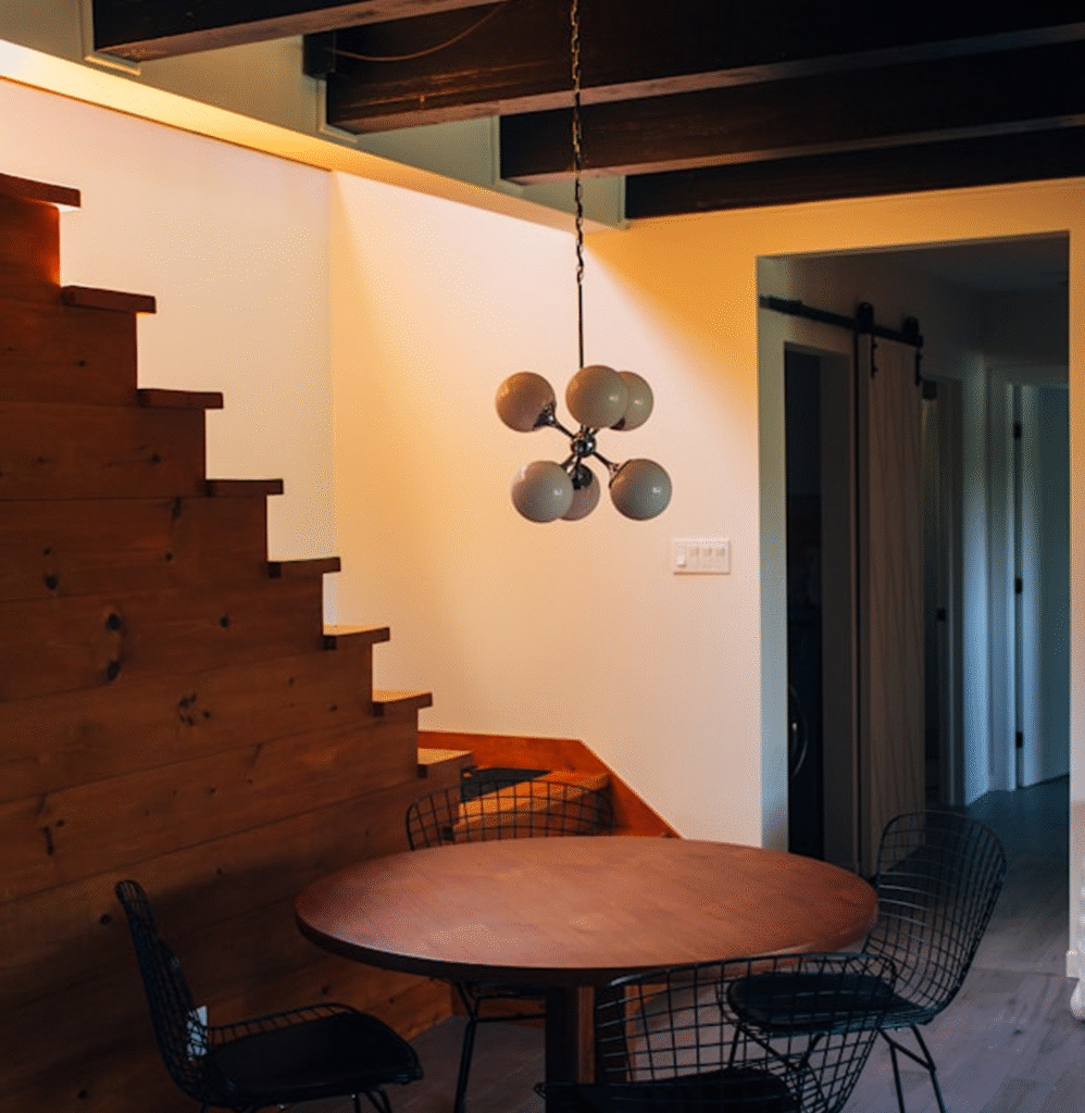 Modern dining area featuring a round wooden table surrounded by black wire chairs, illuminated by a stylish chandelier, with a wooden staircase in the background and a softly lit wall, reflecting Bison Works' home improvement craftsmanship.