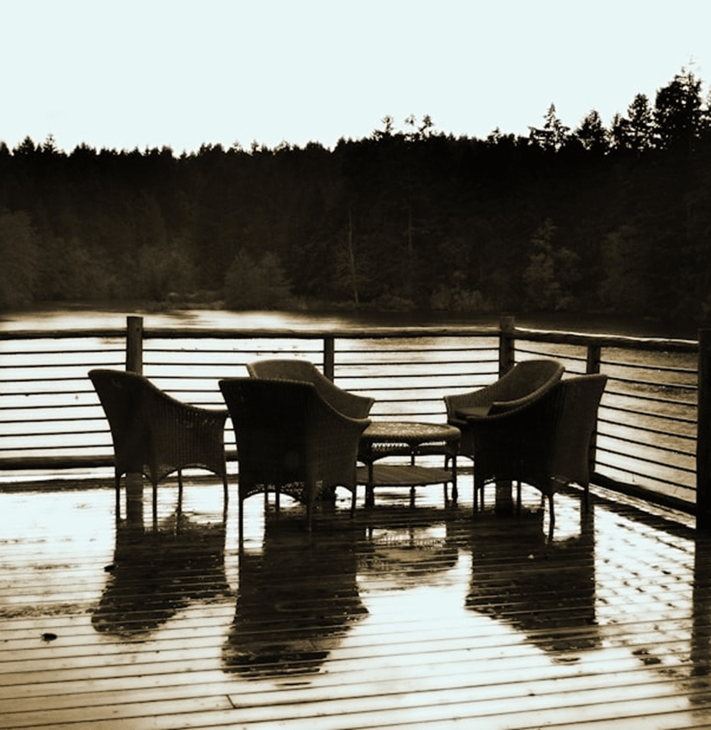 Seating area with wicker chairs and a table on a wet wooden deck overlooking a tranquil lake and forest, highlighting outdoor living space potential for home improvement projects.