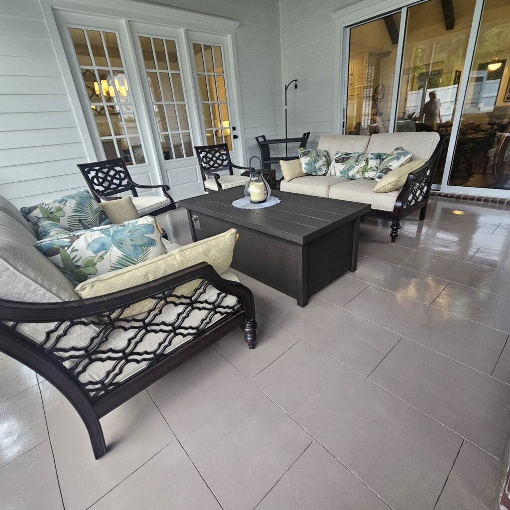 Cozy outdoor seating area with elegant furniture, including a sofa and armchairs adorned with tropical-patterned cushions, complemented by a dark wooden coffee table, set against a backdrop of large glass doors leading indoors.
