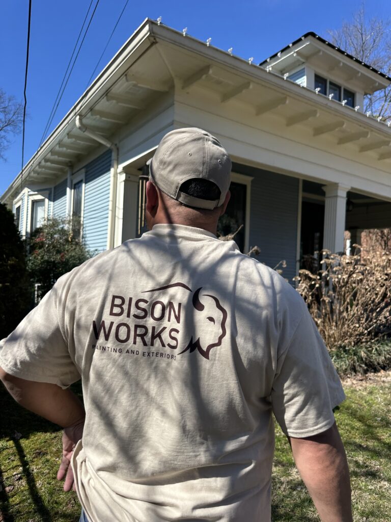Person wearing a Bison Works shirt standing in front of a residential house, showcasing home improvement services related to painting and exterior work in a sunny outdoor setting.