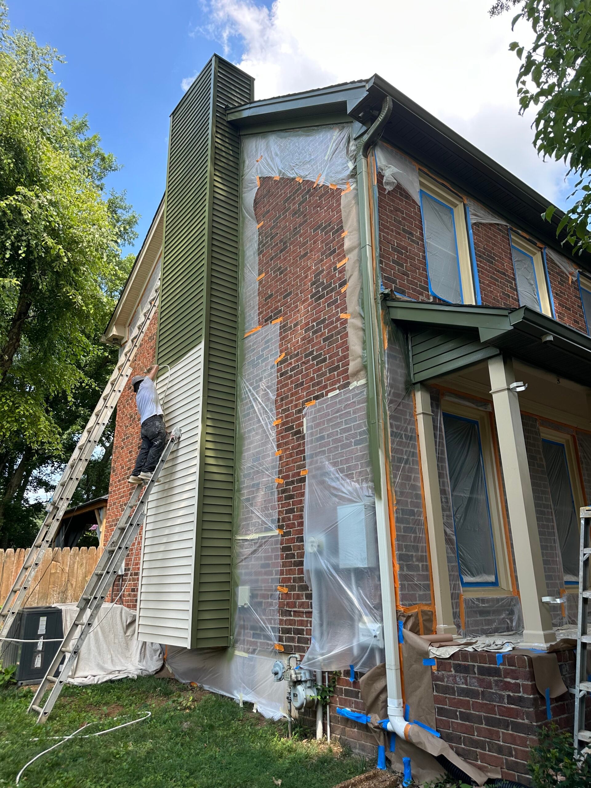 Worker installing green siding on a brick home, with protective plastic covering nearby areas, showcasing home improvement and siding services.