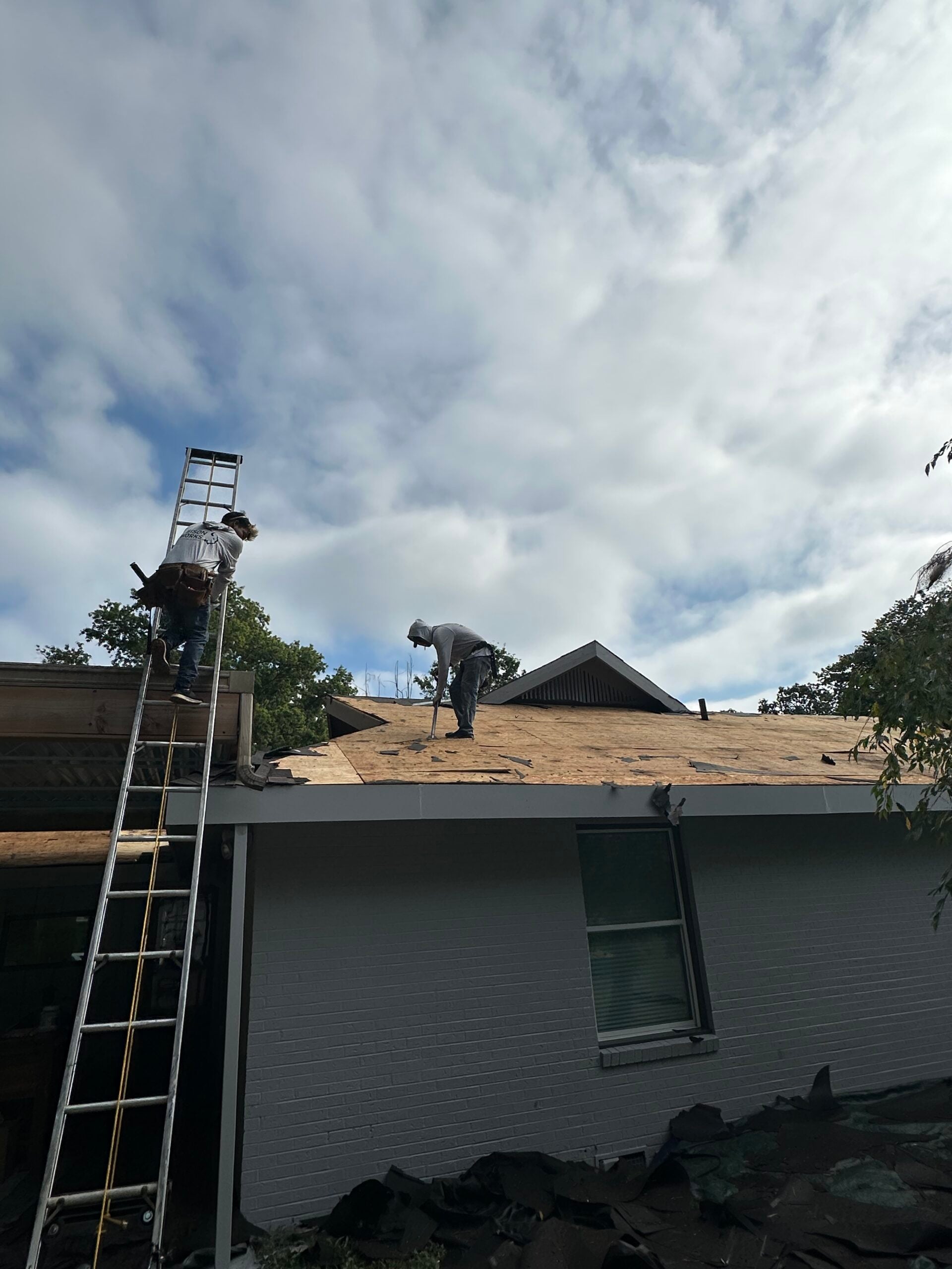 Roofing professionals replacing shingles on a residential roof, with a ladder propped against the house and tools visible on the roof, under a partly cloudy sky.